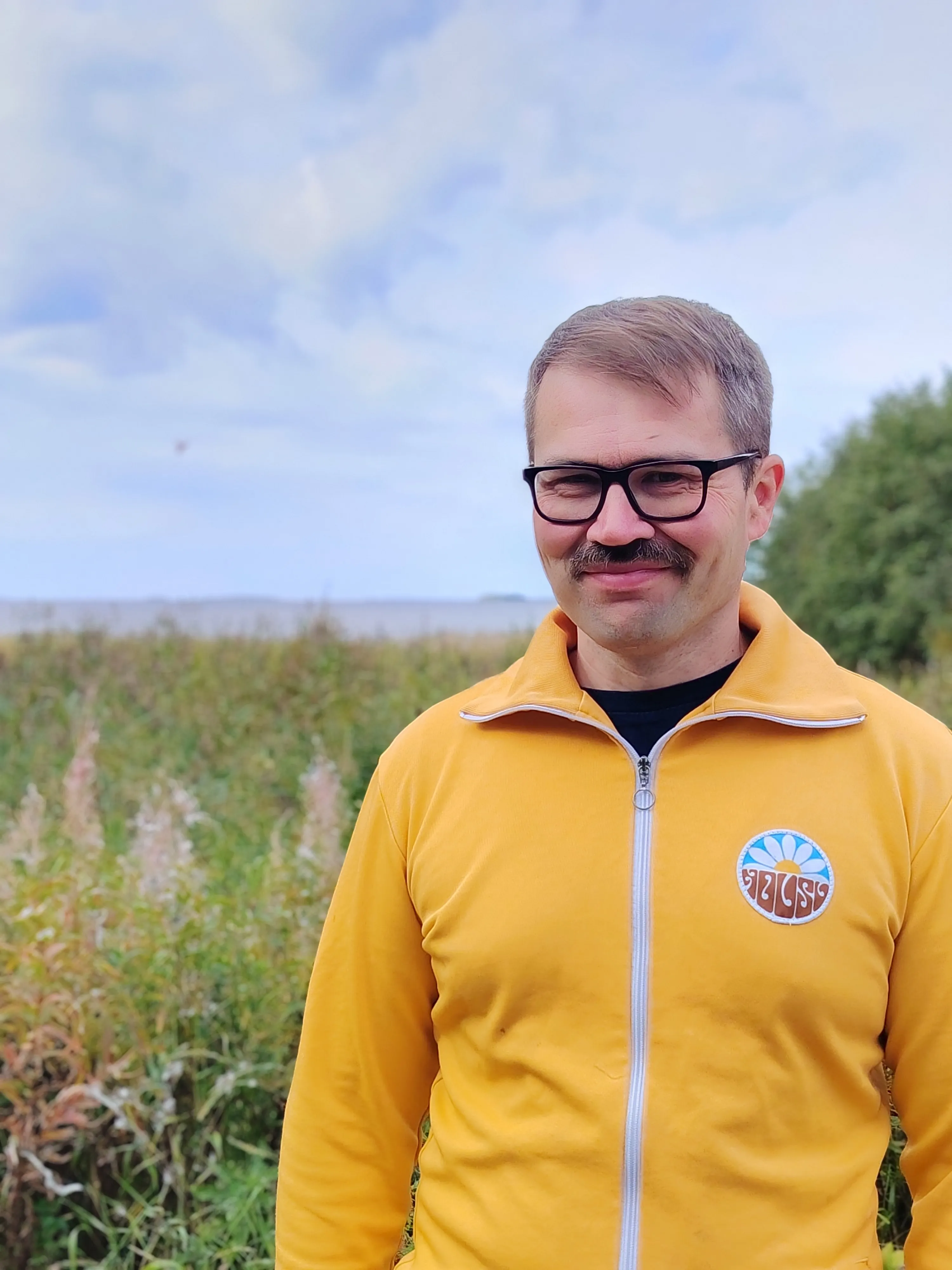 Olli Kohonen hymyilee keltaisessa verryttelytakissa Perämeren rannalla. Taustalla on maitohorsmaa ja merta.
