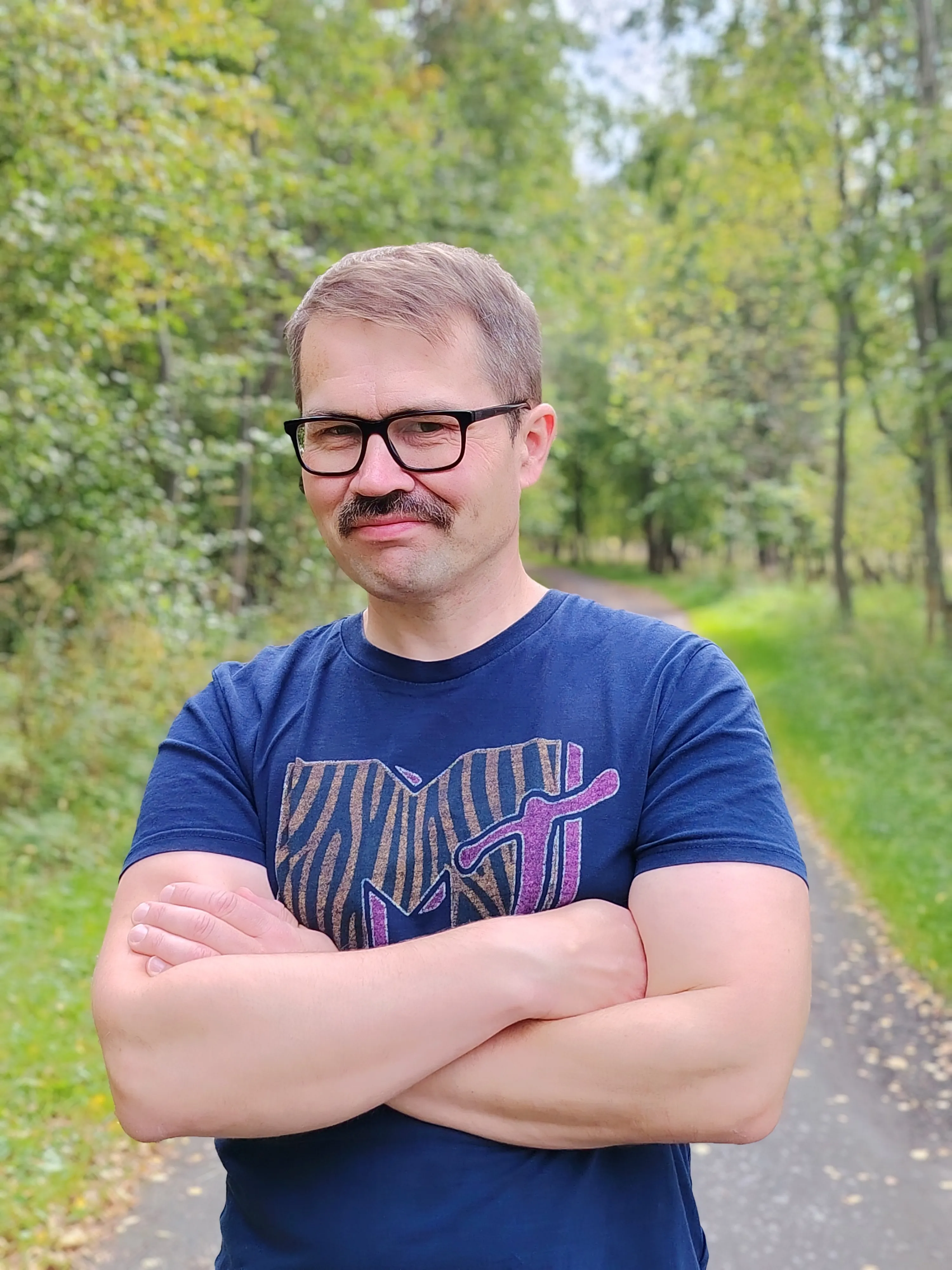 Olli Kohonen seisoo kädet ristissä metsäpolulla sinisessä t-paidassa.